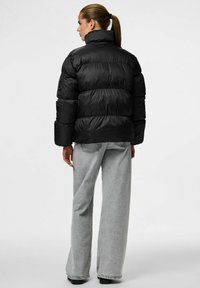 Chaqueta acolchada negra con cuello alto, que presenta secciones acolchadas y puños elásticos, combinada con pantalones de mezclilla de pierna ancha en gris claro.