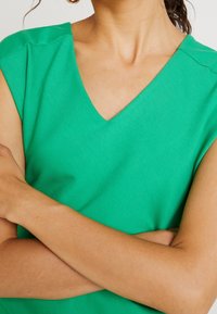 Personne bras croisés portant un haut vert sans manches à col en V, montrant le cou et partiellement les épaules sur un fond neutre.