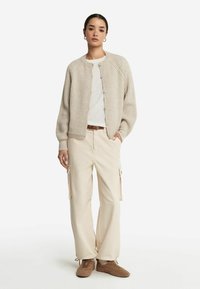 Cardigan beige à côtes avec encolure ronde et fermeture à boutons, porté sur une chemise blanche, associé à un pantalon cargo crème et des chaussons texturés marron.