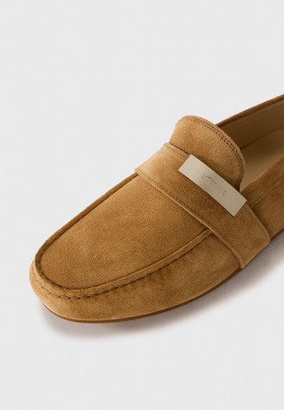 Mocasini din suede bej, cu detalii brodate, o parte superioară netedă și un accent metalic cu brand. Au vârful rotunjit și o talpă flexibilă.