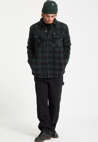 Green and black checkered flannel shirt with two chest pockets, worn over a black shirt, paired with black pants and black shoes.
