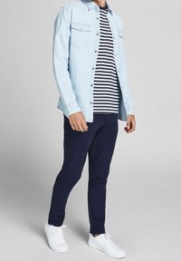 Man i ljusblå denimskjorta över en marinblå och vit randig t-shirt, marinblå byxor och vita sneakers som står mot en grå bakgrund.