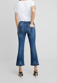Jeans acampanados azules con una cintura de altura media, cinco bolsillos y un dobladillo sin terminar, combinados con una blusa blanca de manga corta y corte holgado, y tacones negros.