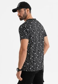 Camiseta negra de manga corta con patrones de texto vertical en blanco. Corte casual, cuello redondo, textura suave, combinada con una gorra negra y jeans.