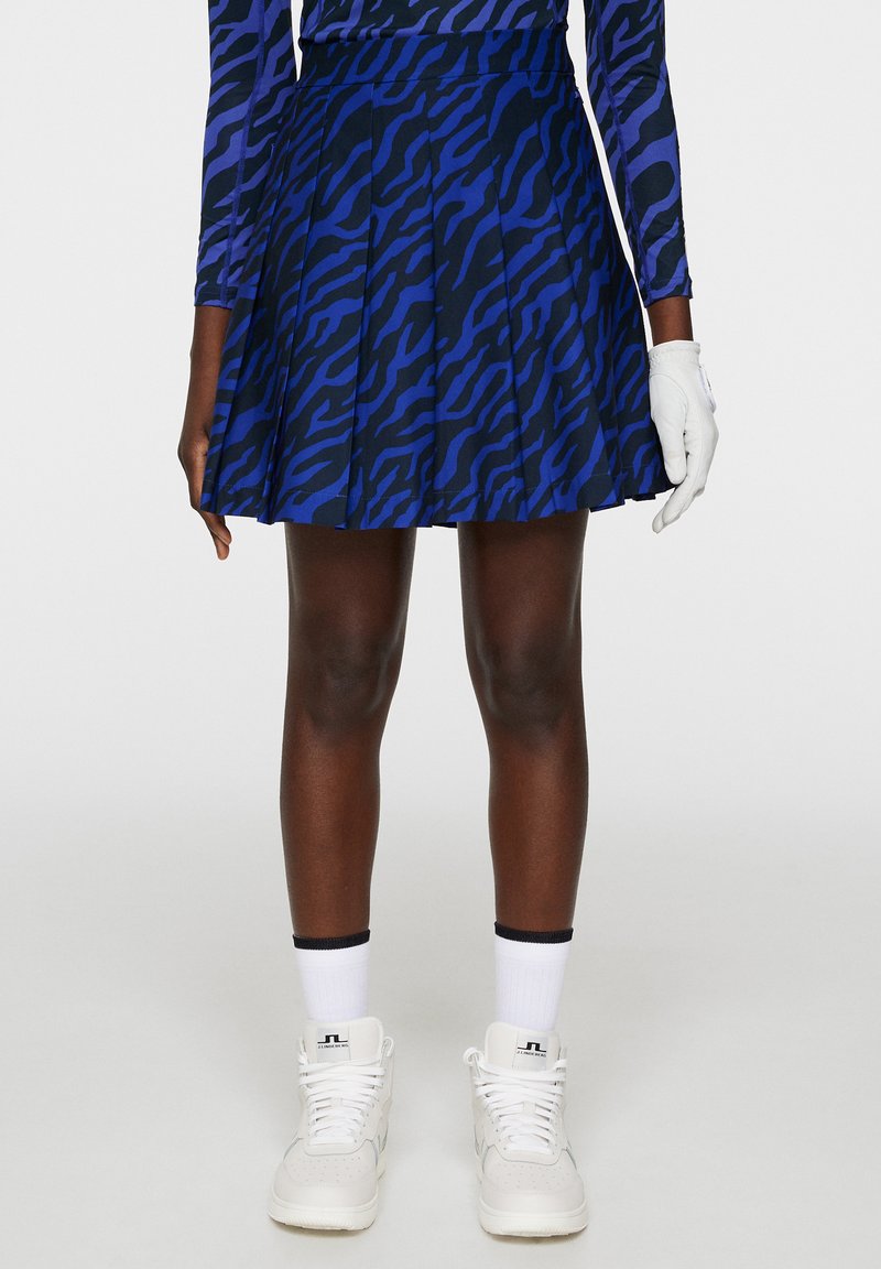 Gonna plissettata blu con un motivo a strisce zebrate nere, realizzata in un tessuto dalla texture liscia. Indossata con sneakers bianche e un guanto.