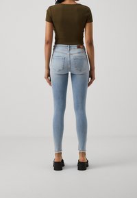 Mujer de pie de espaldas, vestida con jeans ajustados de color azul claro, una camiseta de manga corta verde olivo y zapatos de tacón negro sobre un fondo liso.