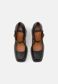 Chaussures Mary Jane en cuir noir avec bouts carrés, boucles de bride réglables et semelles intérieures beige sur fond blanc.