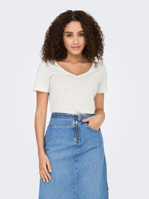 Femme aux cheveux bouclés portant un t-shirt blanc à col en V rentré dans une jupe en jean bleue taille haute avec une fermeture éclair à l'avant et la main dans la poche.