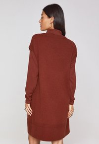 Abito lavorato a maglia di colore ruggine con colletto alto, maniche lunghe, taglio rilassato e dettagli a coste all'orlo e sulle spalle.