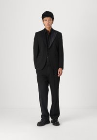 Black suit with a satin lapel, paired with a black button-up shirt and black trousers. Smooth fabric, structured fit, and sturdy black shoes.