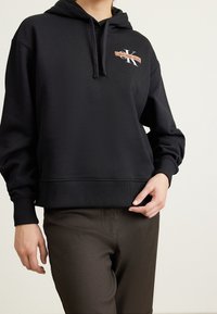 Persoon draagt een zwarte Calvin Klein Jeans hoodie en donkere broek, houdt de zoom van de hoodie met één hand vast tegen een effen achtergrond.