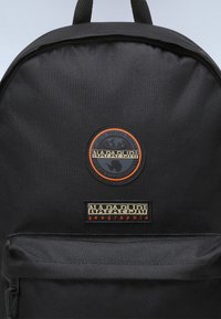 Svart ryggsäck tillverkad av texturerad tyg med runda och rektangulära logotyper i orange och beige på framsidan. Klassisk design med en dragkedjeficka.