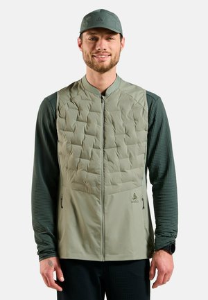 Man wearing olive green quilted vest over long-sleeve dark green shirt and matching olive green cap, standing and smiling.