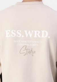 Beige huppari, jossa on suuri valkoinen teksti selässä: "ESS.WRD." yllä "SINCE NINETEEN NINETY ESSENTIALS DEPT." ja "Studio." harmaana.