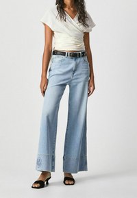 Jasny top w stylu wrap z szerokimi, jasnoniebieskimi dżinsami. Dżinsy mają ozdobne przeszycia oraz logo na krawędzi. Czarne sandały na obcasie z odkrytą toes.