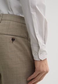 Camicia bianca con piccoli pois marroni, dotata di polsini doppi e bottoni bianchi. Abbinata a pantaloni amaroni chiari con texture.