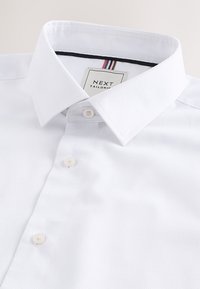 Chemise blanche texturée avec col pointu et patte de boutonnage, montrant une étiquette avec "NEXT Tailoring" à l'intérieur du col.