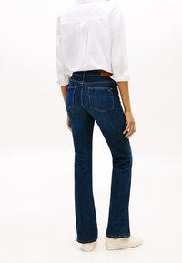 Personne portant un jean bootcut bleu foncé, une chemise blanche à manches longues retroussées et des baskets blanches, debout dos à nous sur un fond uni.