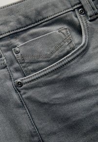 Jeanșii din denim gri au un buzunar din spate cu nasturi negri și cusături contrastante. Textura este netedă, cu un aspect uzat.