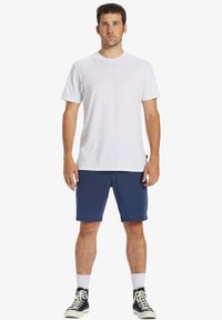 Weißes T-Shirt mit kurzen Ärmeln, blaue Shorts und schwarze High-Top-Sneaker mit weißen Sohlen. Das Modell steht aufrecht mit kurzen Haaren und heller Haut.