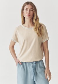 Lyhythihainen, vaaleanbeige t-paita, jossa on pyöreä kaula-aukko ja hienovarainen sininen ja valkoinen raitadetaili hihoissa. Pehmeä kangas, rento malli.