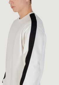 Joven vestido con un sudadera blanca con una franja vertical negra en la manga que presenta una marca Armani Exchange en relieve.