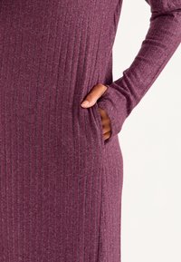Langes, geripptes Kleid in einem tiefen Pflaumenfarbton mit grauen Akzenten. Hat lange Ärmel und seitliche Taschen für zusätzliche Funktionalität.
