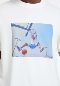 Witte T-shirt met een basketballer in de lucht, gekleed in een blauwe outfit en schoenen, met een bedrukte afbeelding boven een basketbalveld.