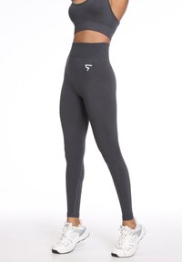 Legginsy sportowe z wysokim stanem w ciemnoszarym kolorze wykonane z gładkiego materiału, z małym białym logo z przodu, w połączeniu z białymi trampkami.