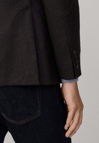Blazer di lana scura con dettagli sui polsini abbottonati, abbinato a un polsino di camicia blu. Sullo sfondo, jeans di denim scuro con cuciture sottili.
