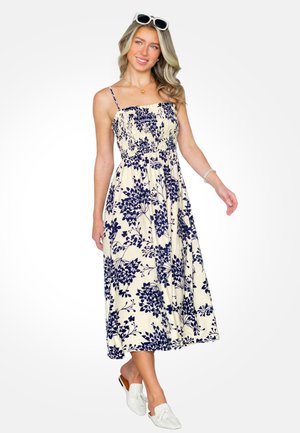 Kobieta o długich blond włosach, nosząca okulary przeciwsłoneczne na głowie i kremową sukienkę midi z niebieskim kwiatowym wzorem, do tego białe buty wsuwane.