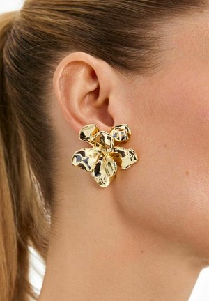 Zelta auskars zieda formā ar teksturētu virsmu, kas izceļ spožu apdari un slāņotu ziedlapu dizainu, valkāts modeļa ausī.