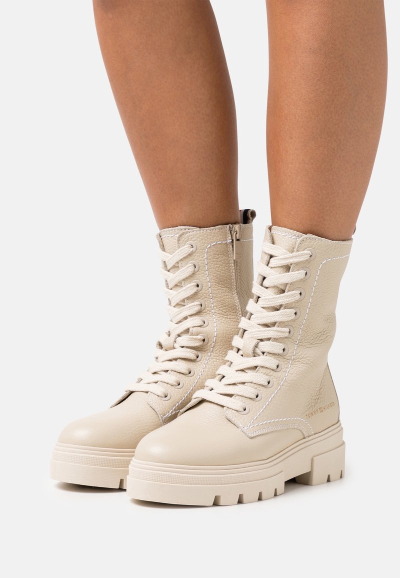 Bottes en cuir beige à lacets avec semelles épaisses portées sur des jambes nues, dotées de fermetures éclair latérales et de détails de couture blanche.