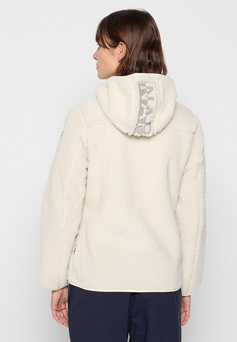 Femme portant une veste en sherpa crème avec capuche, ornée d'un logo gris vertical, vue de dos.