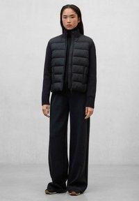 Chaleco acolchado negro con cuello alto, cierre de cremallera y diseño acolchado, sobre una camiseta negra de manga larga y pantalones negros de pierna ancha.