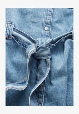 Jeansipaita, mis on helesinise värvusega, nupuga kinnitused ja seotud detail talje piirkonnas, nähtava õmbluse ja tekstuuriga.