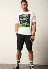 Camiseta de algodón blanca con un gráfico negro de la cara de un payaso y texto verde "EL CIRCUS DE LOS PECADORES", combinada con pantalones cortos de carga negros y zapatillas negras.