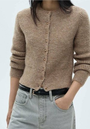 Femme portant un cardigan en tricot côtelé beige avec des boutons en perle et un jean taille haute gris clair avec une ceinture noire.