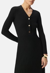 Abito a maglia ribbed nero con scollatura a V, maniche lunghe e bottoni dorati lungo la parte anteriore. Presenta una vita aderente e una gonna fluente.