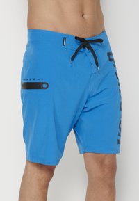 Bañador azul con cordón negro, que cuenta con un bolsillo lateral y letras negras verticales en la pierna izquierda. Tejido suave y ligero.