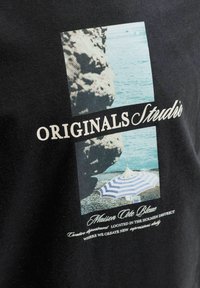 Βραχώδης ακτή και γαλάζια θάλασσα πάνω από μια παραλία με βότσαλα και μια μπλε και άσπρη ριγέ ομπρέλα, κείμενο "ORIGINALS Studios" και γαλλικές φράσεις παρακάτω.