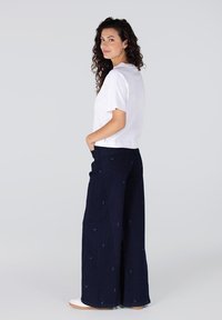 Witte top met korte mouwen en marineblauwe brede broek met geborduurde details. Model staat met handen in de zakken en draagt witte schoenen.