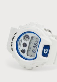 Montre G-SHOCK blanche avec un accent bleu autour de l'affichage numérique, dotée de boutons, d'un bracelet texturé et d'une conception résistante aux chocs.
