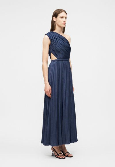 Femme debout portant une robe maxi plissée bleu marine asymétrique à une épaule, sans manches, avec une découpe sur le côté, et des sandales à talons noires.