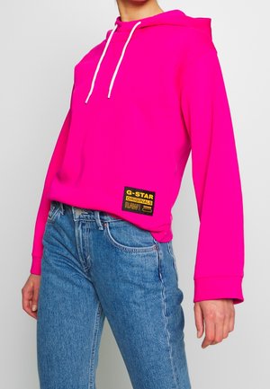 Osoba w jaskraworóżowej bluzie z białymi sznurkami i niebieskich dżinsach, pokazująca metkę G-Star Originals RAW denim z przodu bluzy.