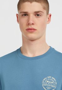 T-shirt en coton bleu avec un col rond, arborant un logo circulaire blanc sur le côté gauche de la poitrine. Texture lisse, coupe décontractée.