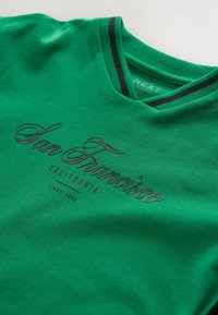 T-shirt in cotone verde con scollo a V, caratterizzato da dettagli a strisce nere sul colletto e testo in rilievo che dice "San Francisco, California, dal 1982."