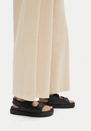 Czarne sandały na rzepy, z wytrzymałą podeszwą i odkrytymi palcami, noszone z jasnobeżowymi szerokimi spodniami.