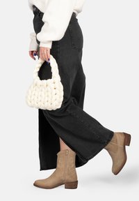 Borsa intrecciata bianca con manico curvo, abbinata a jeans a gamba larga neri e stivaletti beige con cuciture decorative.