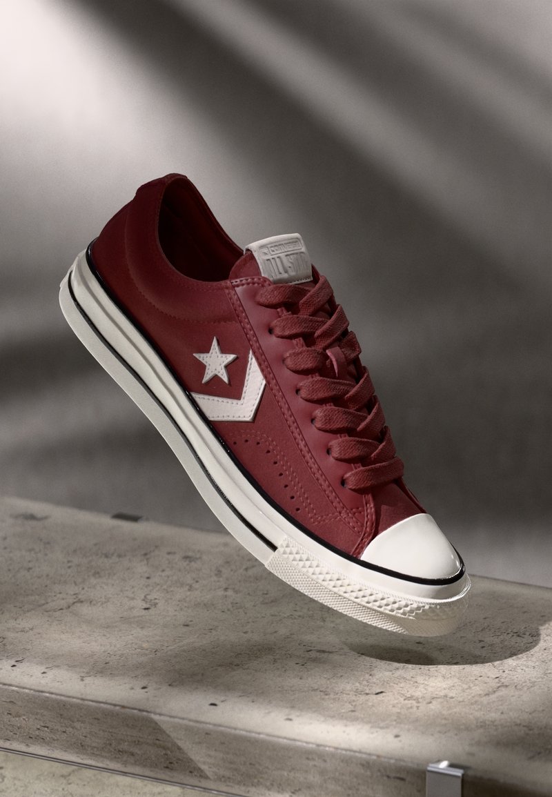 Baskets basses rouges avec logo étoile blanc, bout blanc et semelle rayée noire flottant au-dessus d'une surface en béton.